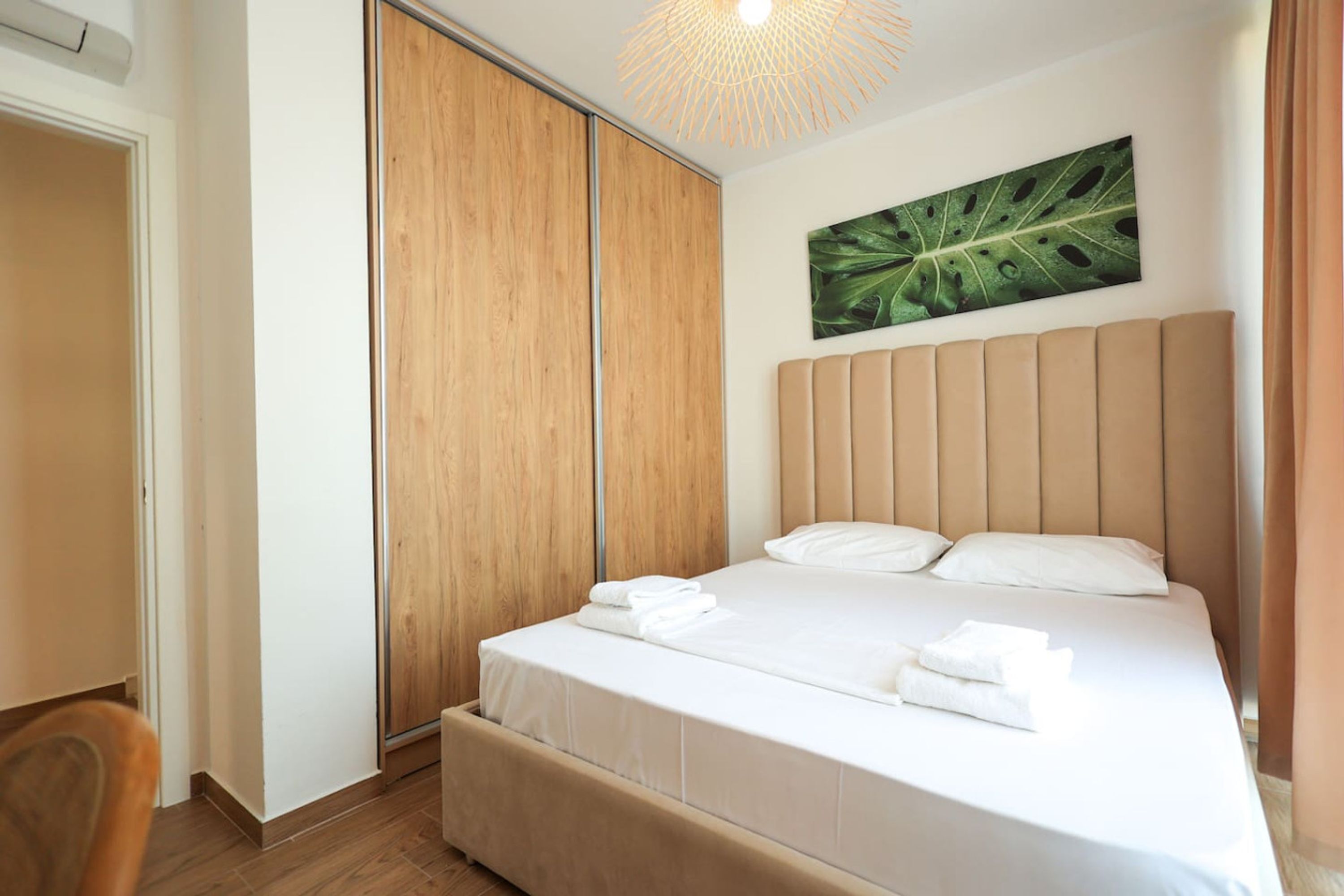 Een opgemaakt bed met witte lakens en handdoeken, een houten garderobe, en een groot kunstwerk met bladeren aan de muur.