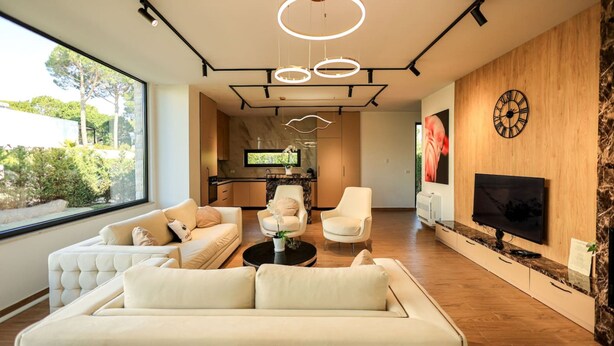 Een moderne woonkamer met een bank, fauteuils, een salontafel en een aan de muur bevestigde televisie. Er is een houten accentmuur en een groot raam met uitzicht op groen buiten.