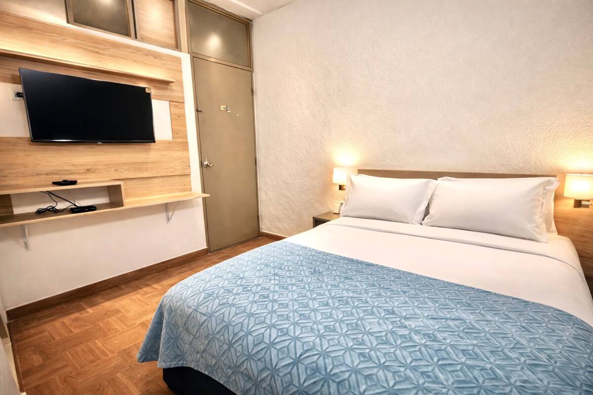 Habitación de hotel con cama, televisor de pantalla plana montado en la pared, armario y mesita de noche con lámpara.