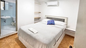 Ein Hotelzimmer mit Bett, Nachttisch, Wand-Klimaanlage und einem durch eine Glastür einsehbaren Badezimmer.