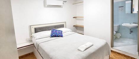 A hotel room with a bed, a bathroom with a shower, and a white air conditioning unit on the wall.