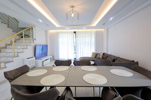A modern living room with a dining area, featuring a staircase with golden railings, a flat-screen TV, and a large window with sheer curtains.