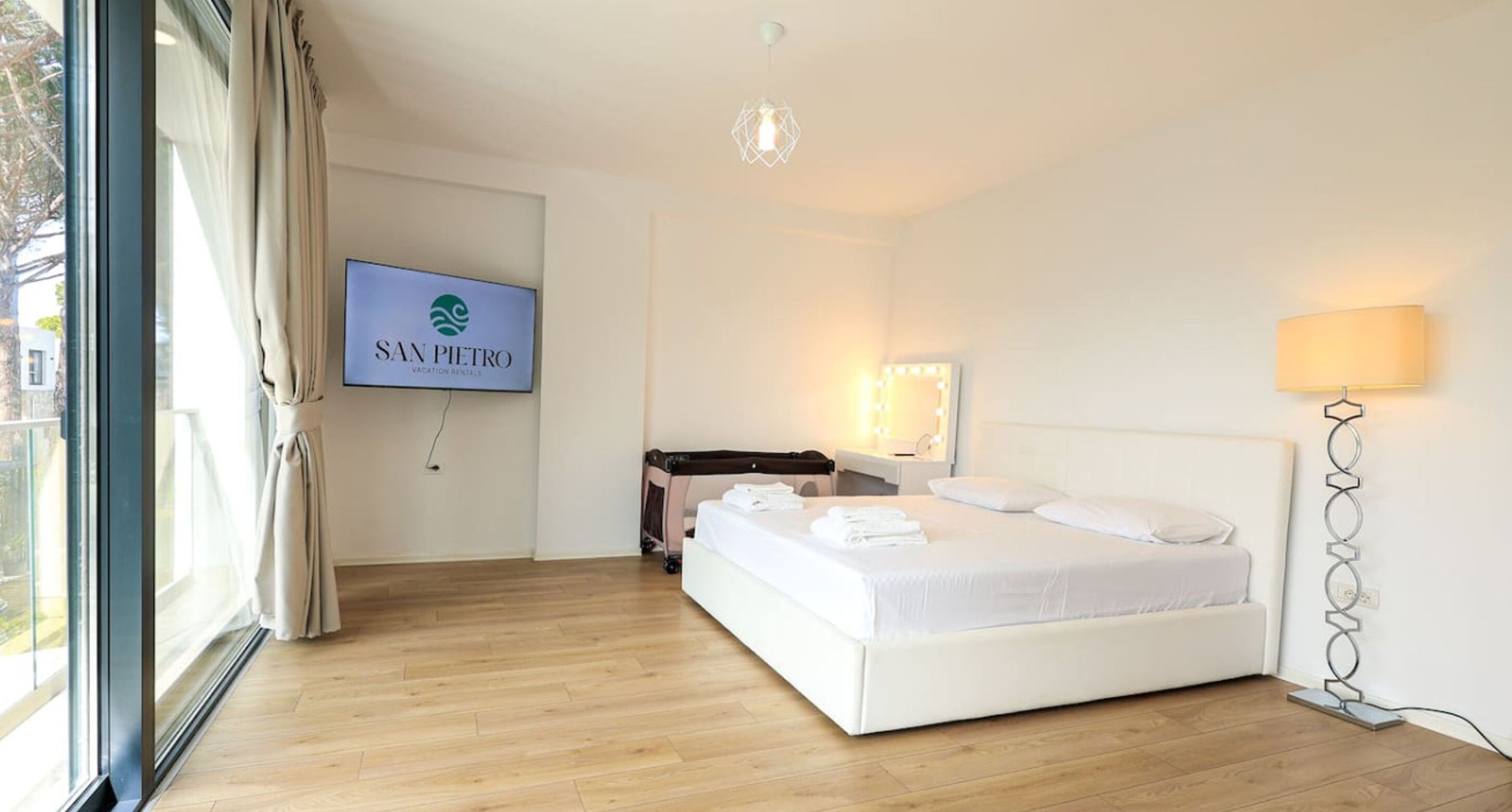 A hotel room with a bed, a TV, a bedside table, a lamp, and a view of the outdoors.