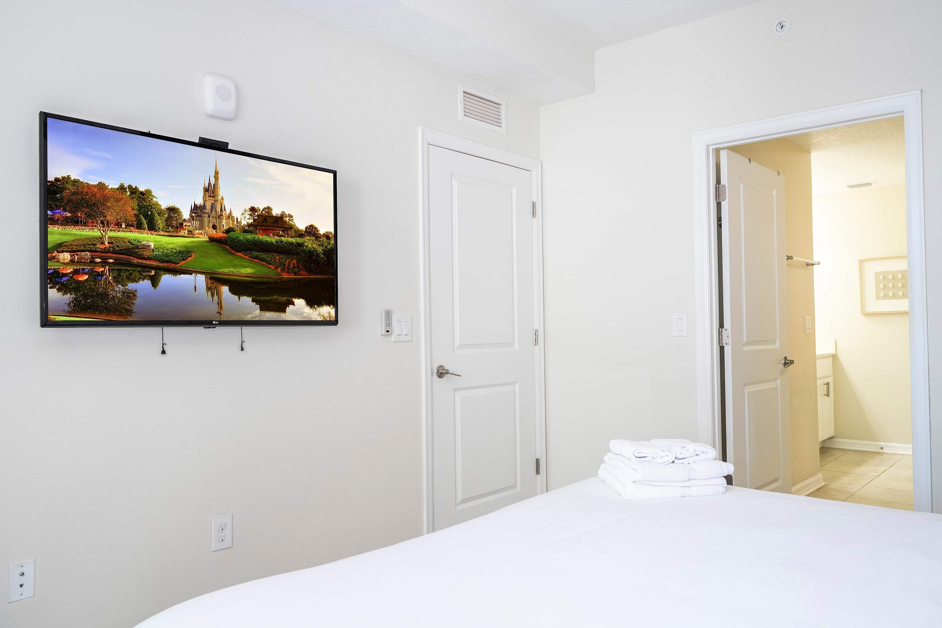 Habitación de hotel con televisor de pantalla plana en la pared mostrando un paisaje, cama con ropa blanca bien tendida y una puerta cerrada a la derecha.