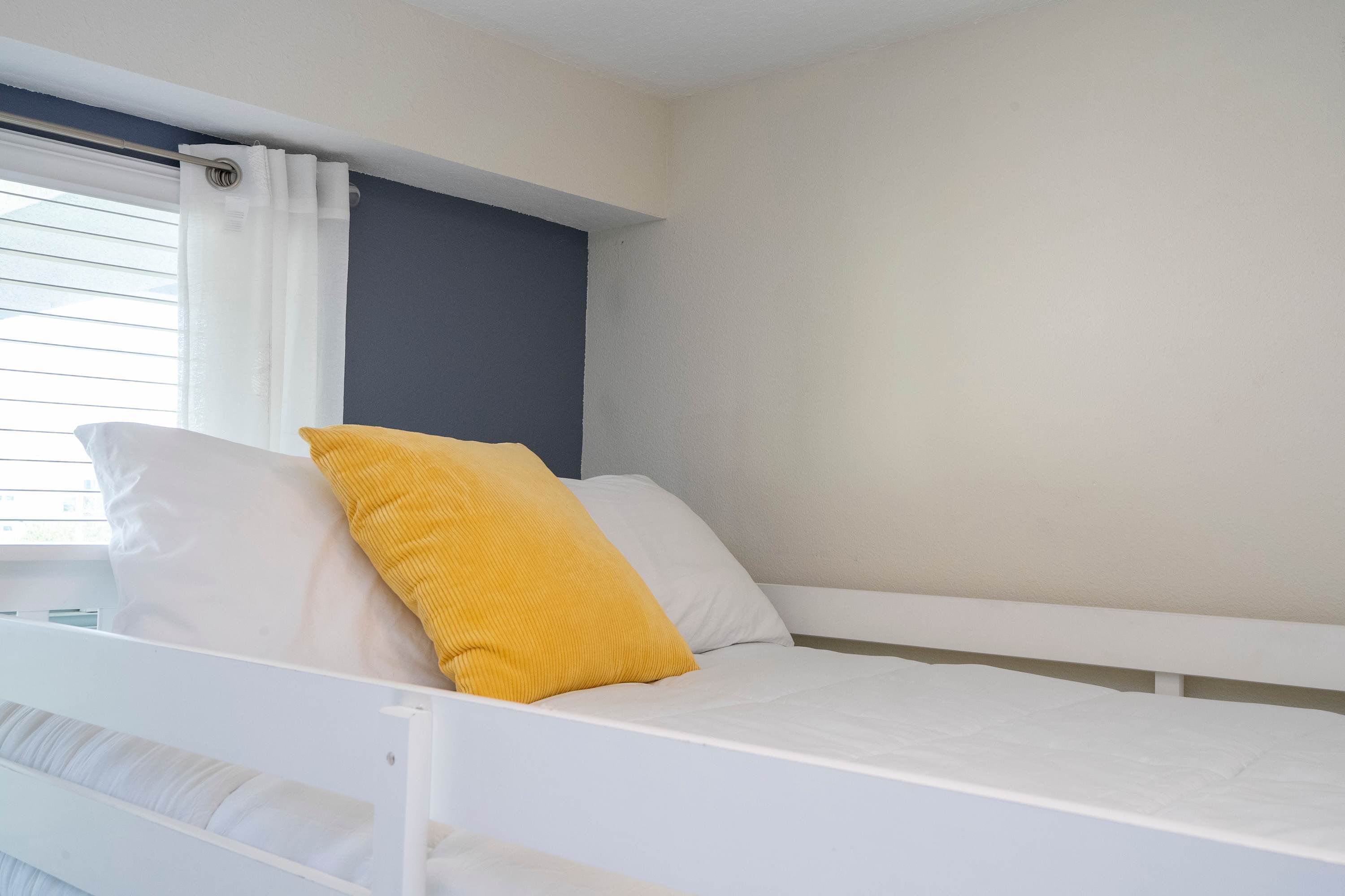 Uma cama de solteiro com roupa de cama branca e travesseiro amarelo, uma janela com cortinas brancas e uma parede lisa com detalhe escuro.