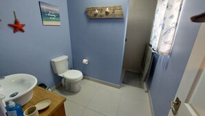 Un baño con inodoro, lavamanos y un estante de madera con artículos de aseo.