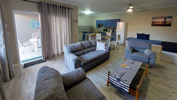 Una sala de estar con sofá, sillón y una mesita. Se ve una cocina al fondo con armarios y un refrigerador.