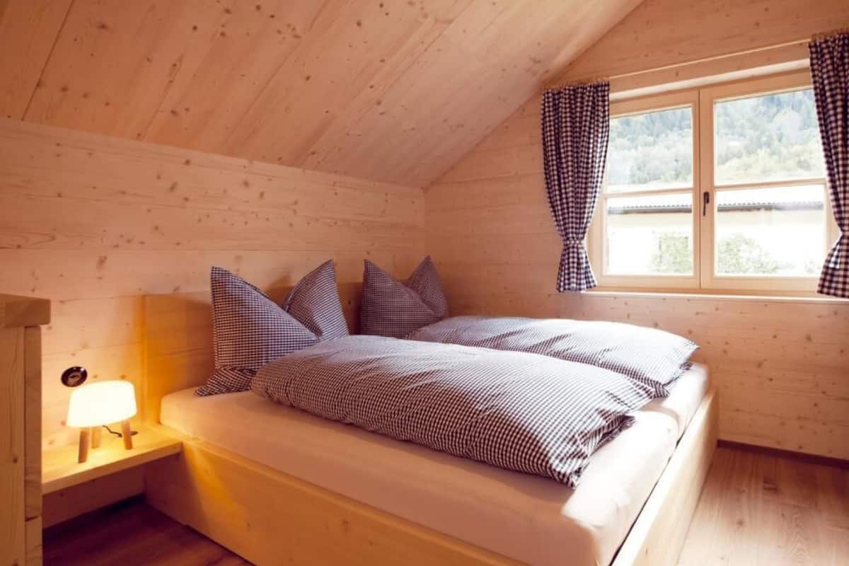 A wooden cabin bedroom with a bed, pillows, a nightstand, a window with curtains, and a view of the outdoors.