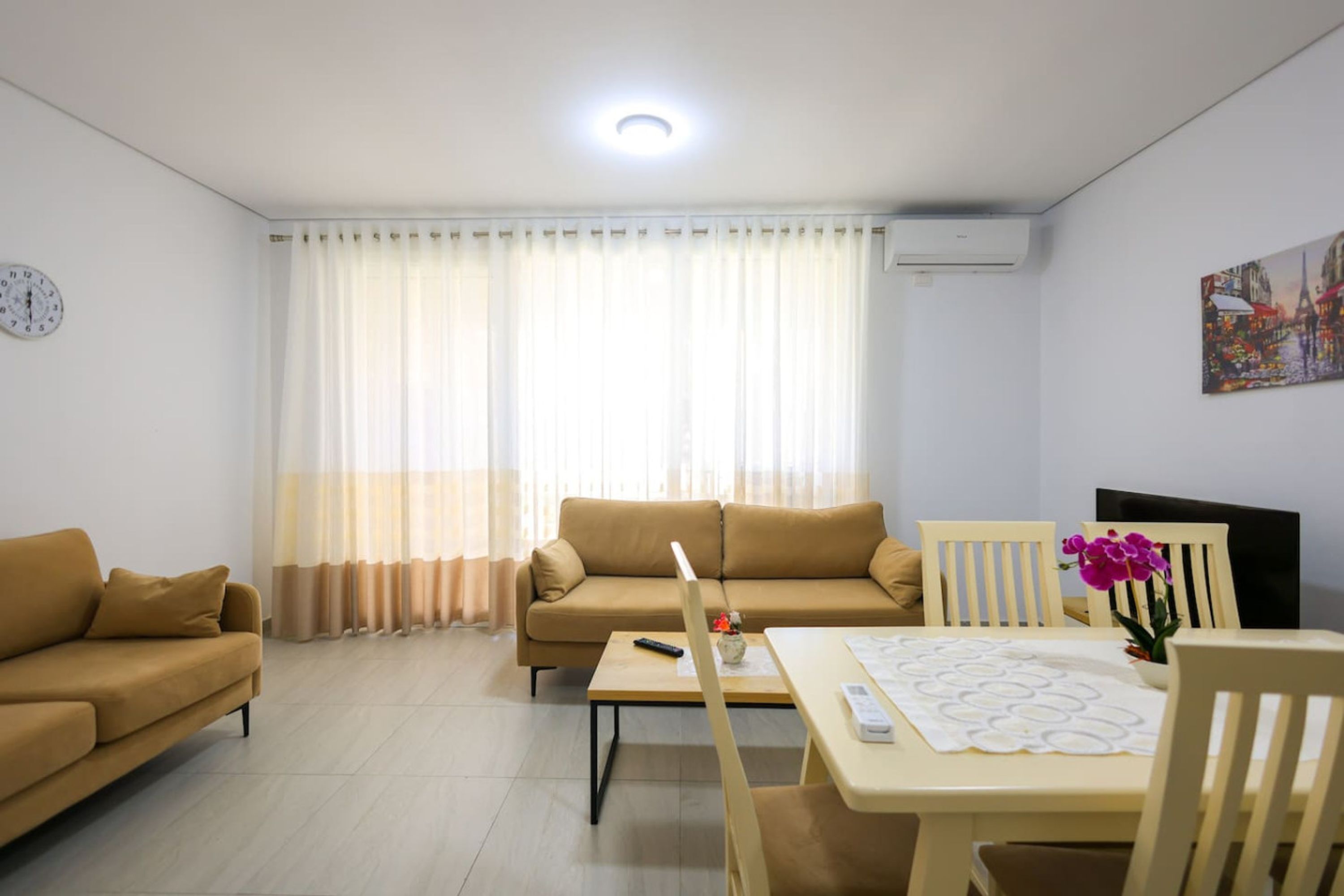Una sala de estar con un sofá, una mesa de comedor con sillas, un reloj en la pared y un ventanal con cortinas.