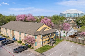 A two-story building with a parking lot, a staircase, and a large stadium in the background. - AT&T at Your Doorstep • 2BR + Parking (Arlington)
