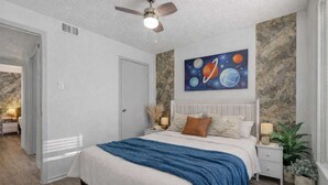 Un dormitorio con cama, mesitas de noche, ventilador de techo, un cuadro de planetas y una pared de piedra.