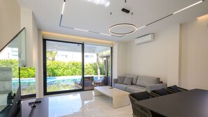 A modern living room with a large glass door leading to a balcony, a grey sofa, a coffee table, and a dining table with chairs.