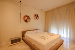 Quarto de hotel moderno com uma cama grande, uma pequena mesa de cabeceira e uma janela com cortinas leves.