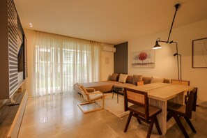 Een moderne woonkamer met een bank, een eettafel en stoelen. Er is een groot raam met dunne gordijnen, een vloerlamp en een schilderij aan de muur.