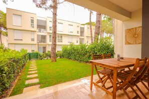Uma área de pátio com mesa e cadeiras de madeira, um pequeno jardim com gramado e árvores, e um edifício com varandas ao fundo.