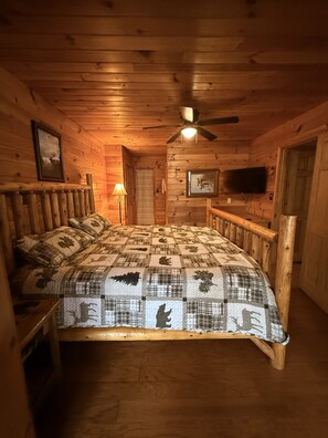 Un dormitorio de cabaña de madera con una cama, un ventilador de techo, un televisor montado y una puerta.