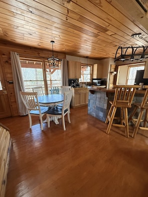 A wooden cabin interior with a dining table, chairs, a kitchen area, and a stone fireplace.