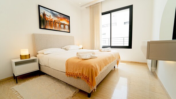 Un hotel moderno con una cama grande, una mesita de noche con lámpara, un ventanal con vistas a edificios y un cuadro en la pared.