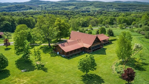 A large, rustic log cabin with a red roof, surrounded by lush green fields and trees, set in a hilly landscape. - Luxurious 4-bedroom cabin that sleeps 13 close to Cooperstown NY with AC (Burlington Flats)