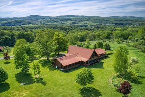 A large, rustic log cabin with a red roof, surrounded by lush green fields and trees, set in a hilly landscape.