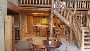 A rustic interior with a wooden dining table and chairs, a kitchen area, and a wooden staircase. - Luxurious 4-bedroom cabin that sleeps 13 close to Cooperstown NY with AC (Burlington Flats)