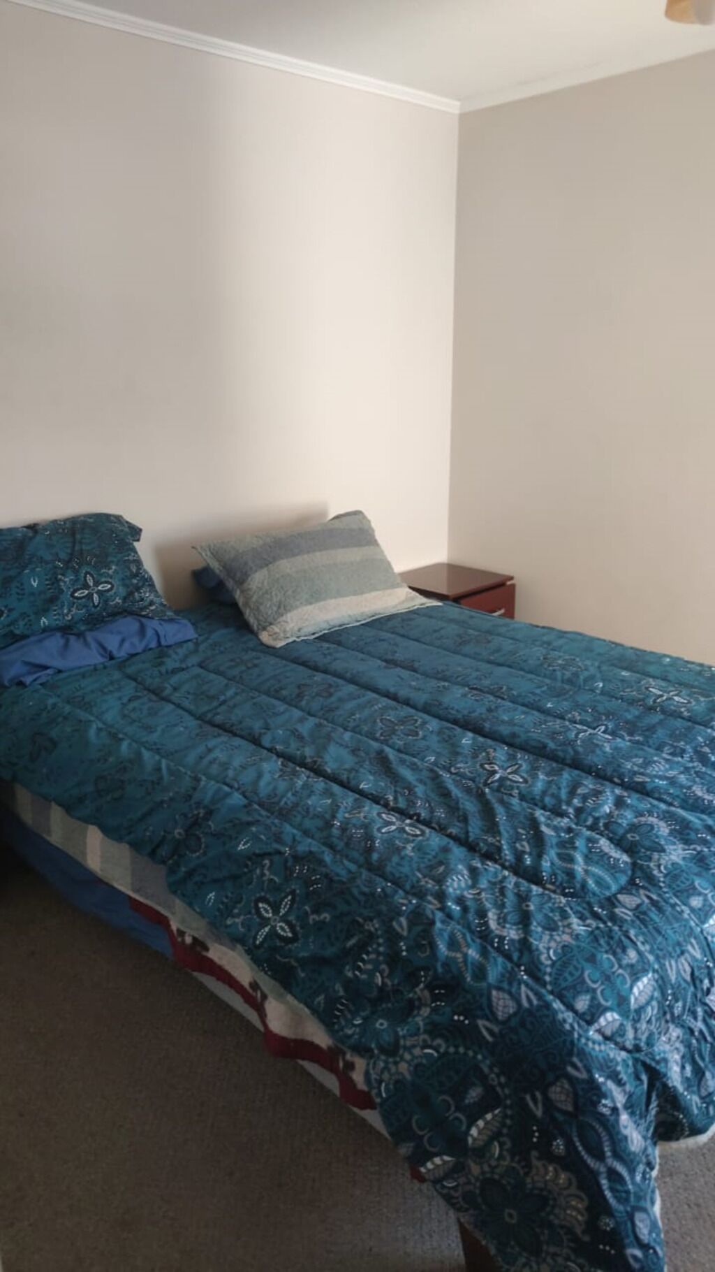 A single bed with a blue patterned bedspread and pillows, a bedside table, and a plain wall.