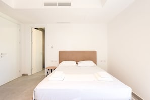 A minimalist hotel room with a large bed, a small side table, and a door leading to another room.