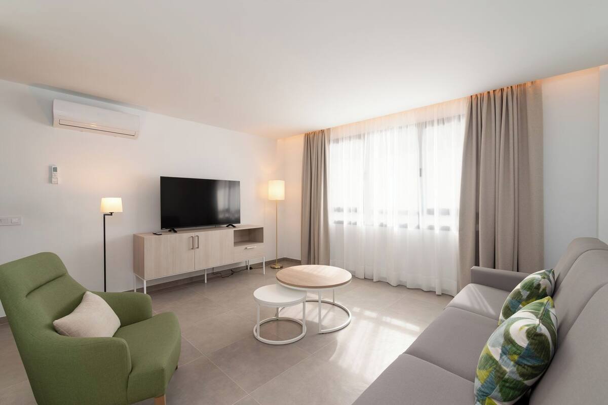 A modern living room with a flat-screen TV, a green armchair, a grey sofa, a small round table, and a floor lamp.