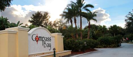 Señal de Compass Bay con una parte superior curva y un emblema de estrella, rodeada de palmeras y un cielo despejado.