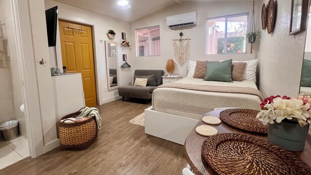 Une chambre à coucher comprenant un lit, des tables de chevet, une commode, une chaise, une fenêtre avec des stores, un appareil de climatisation et une porte.