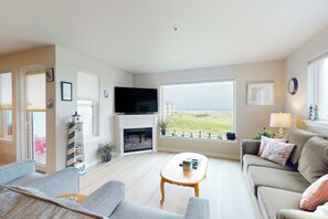 Ein Wohnzimmer mit Kamin, Fernseher, einer Couch, einem Couchtisch und Blick ins Freie.