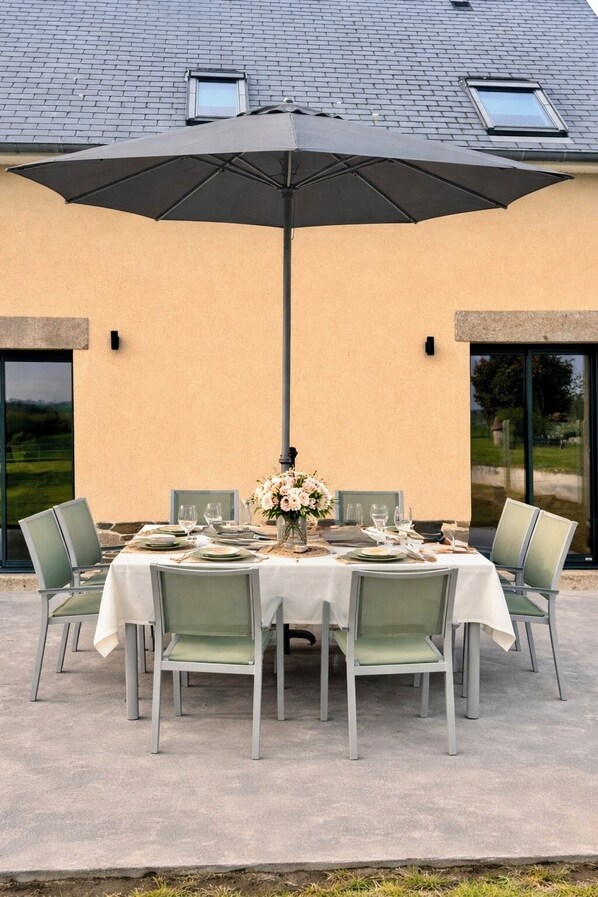 Un espace de restauration en plein air avec une table dressée pour quatre personnes, entourée de chaises, et protégée par un grand parasol.