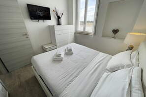 Ein ordentlich bezogenes Bett mit weißen Leinen und Handtüchern, ein an der Wand befestigter Fernseher, eine Kommode, ein Fenster mit Blick und eine herzförmige Dekoration an der Wand.