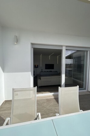 Ein Balkon mit zwei Stühlen, eine Schiebetür zu einem Wohnbereich mit einer Couch und einem Fernseher.
