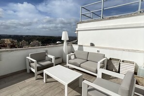 Terraço no topo do prédio com móveis brancos, incluindo sofás e uma mesa de centro, e um abajur grande e branco.