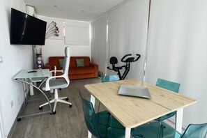 A modern, well-lit room with a wooden table, green chairs, a white sofa, a TV mounted on the wall, and a stationary exercise bike. - Attico con splendida vista su Villa Ada (Roma (RM))