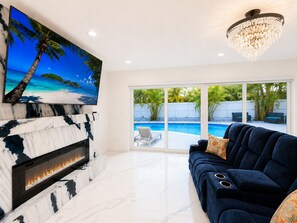 Ein Wohnzimmer mit Kamin, großem Fernseher, einem dunkelblauen Sofa und Blick auf einen Swimmingpool.