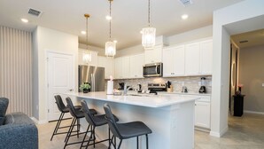 Une cuisine moderne avec des meubles blancs, un îlot central doté de tabourets de bar, des appareils en acier inoxydable et un éclairage suspendu.