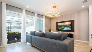 Un salon moderne avec un canapé gris, un téléviseur à écran plat fixé au mur et une grande fenêtre avec des rideaux.