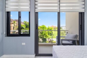 A bedroom with a bed, a balcony with a chair, and a view of greenery and buildings. - Coastal Escape in Riviera, Tower Isle~Beach Nearby (Tower Isle)