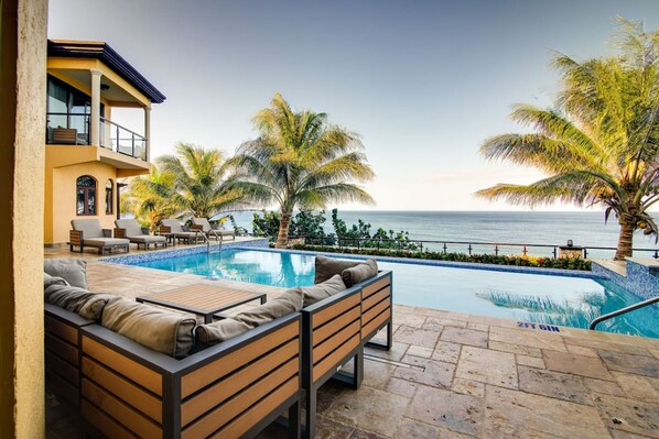 Un espace aménagé au bord de la piscine, avec vue sur l’océan, une terrasse en bois et un coin salon avec des coussins.