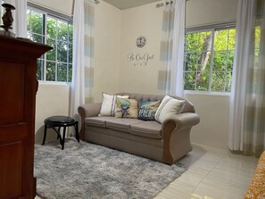 A room with a beige sofa, a small stool, a fluffy rug, and a wall with a 'Be Our Guest' sign. - Adorable 1BR Cottage Walk to the Beach Priory (Priory)