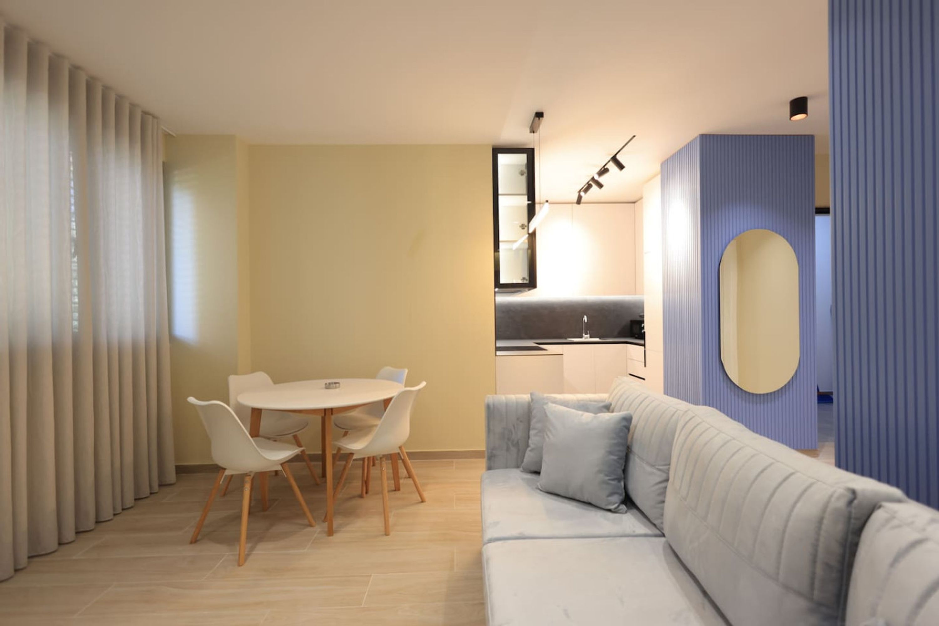 A modern living room with a light-colored sofa, a dining table with chairs, and a kitchen area visible in the background.