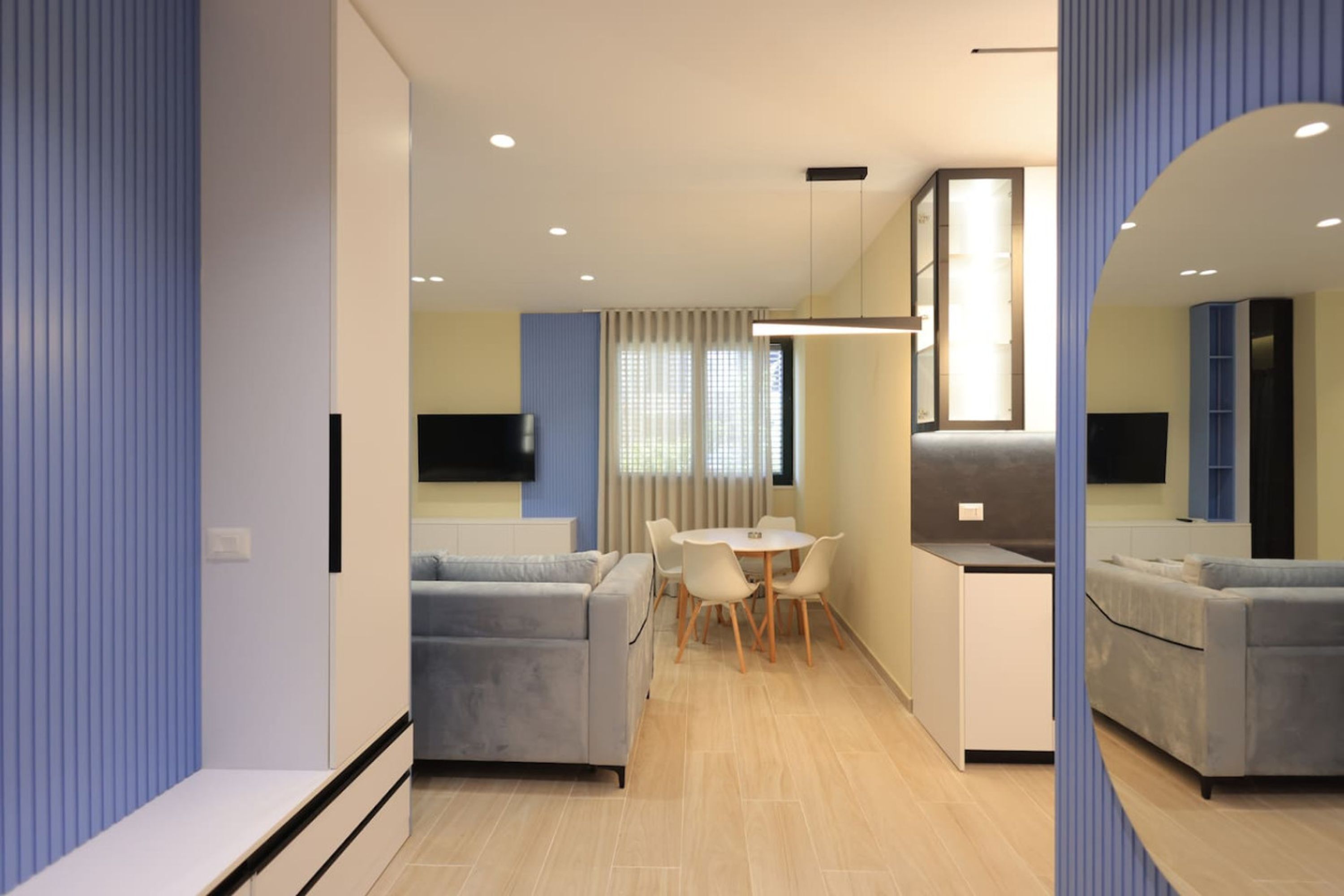 A modern living room with a grey sofa, a small dining table with chairs, and a kitchen area with a microwave and a flat-screen TV.