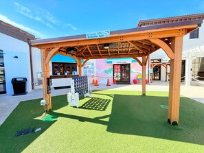 Ein überdachter Außenbereich mit Kunstgrashalm, einer Holzpergola und einem Schild mit der Aufschrift 'Westside Willy's Activities Cove'.