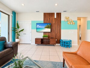 A modern living room with a flat-screen TV, a wooden cabinet, a dining area with blue chairs, and a large window with curtains. - Family Villa in Windsor • Lazy River • BBQ • Pool (Kissimmee)