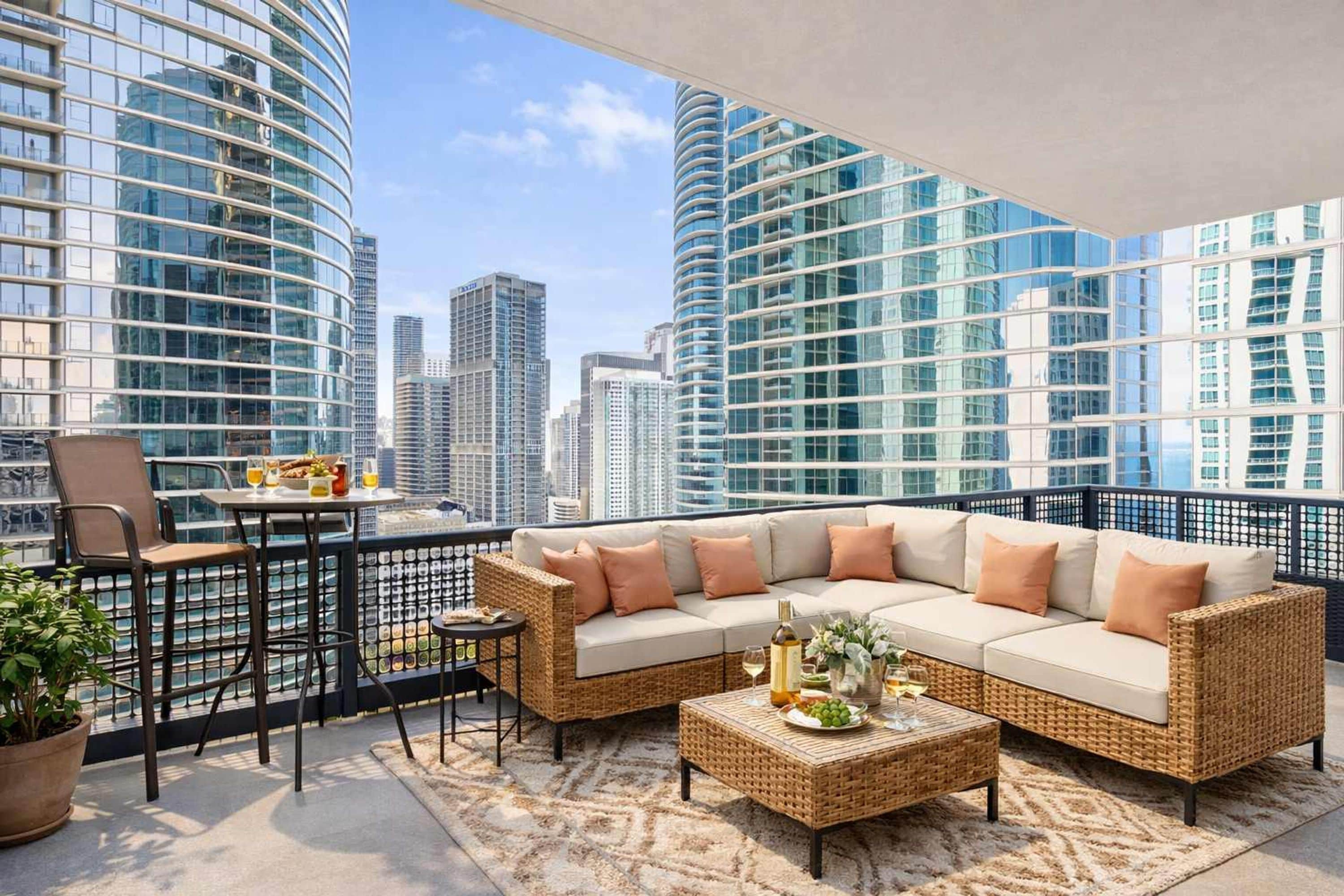 A balcony with a wicker sofa, a small table, and a chair, overlooking a cityscape with tall buildings.