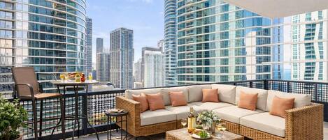 A balcony with a wicker sofa, a small table, and a chair, overlooking a cityscape with tall buildings.