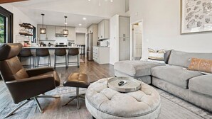 Ein modernes Wohnzimmer mit einer grauen Couch, einem braunen Ledersessel und einem runden Hocker. Im Hintergrund ist eine Küche mit weißen Schränken und ein Essbereich mit Barhocker zu sehen.