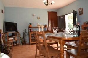 Un comedor con una mesa de madera dispuesta para cuatro personas, rodeada de sillas de madera. A la izquierda, un televisor sobre un mueble bajo, y una puerta que da al exterior.